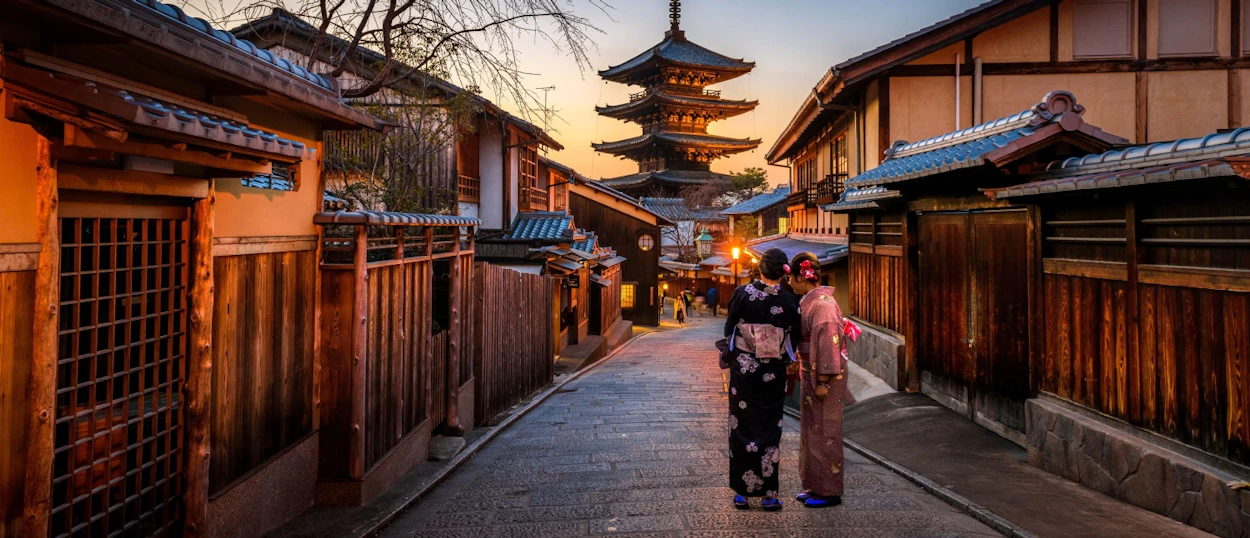 Two people in kimonos walk down a traditional street towards a pagoda at sunset in Kyoto.