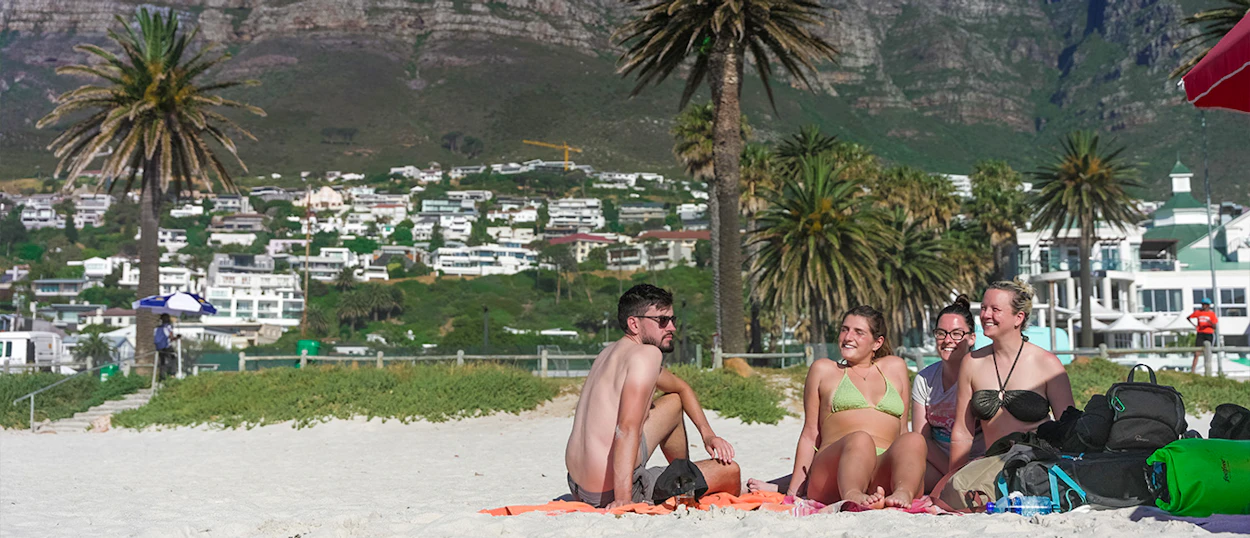 Four friends relax on a sunny beach with palm trees and distant mountains in Cape Town.
