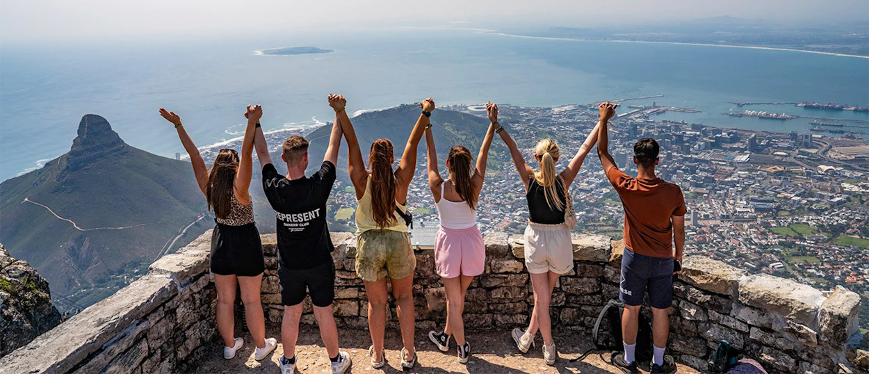 Six friends hold hands, celebrating a panoramic view of Cape Town from Table Mountain.