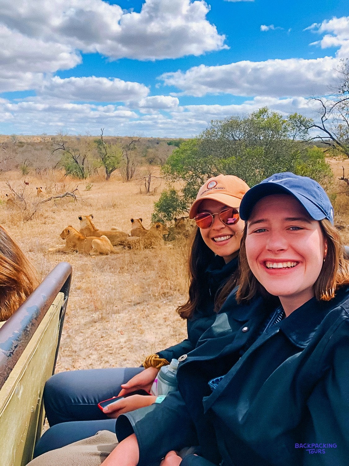 Two travellers grinning for a selfie from a safari vehicle as lionesses rest behind them