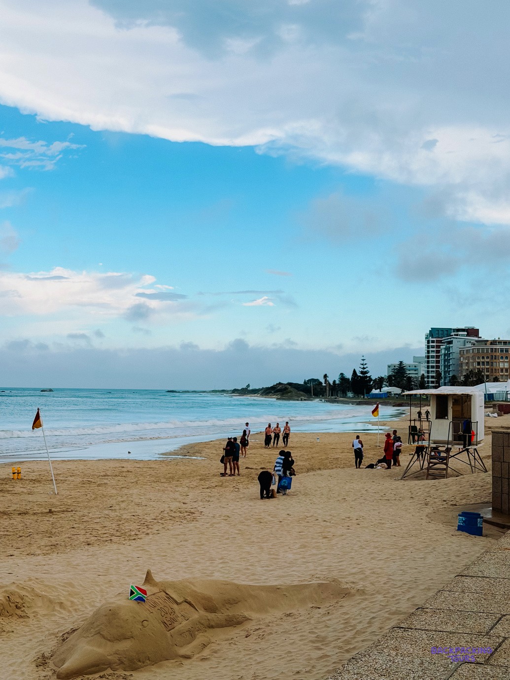 Sandy beach with a lifeguard hut, swimmers and seafront apartment blocks under a blue sky