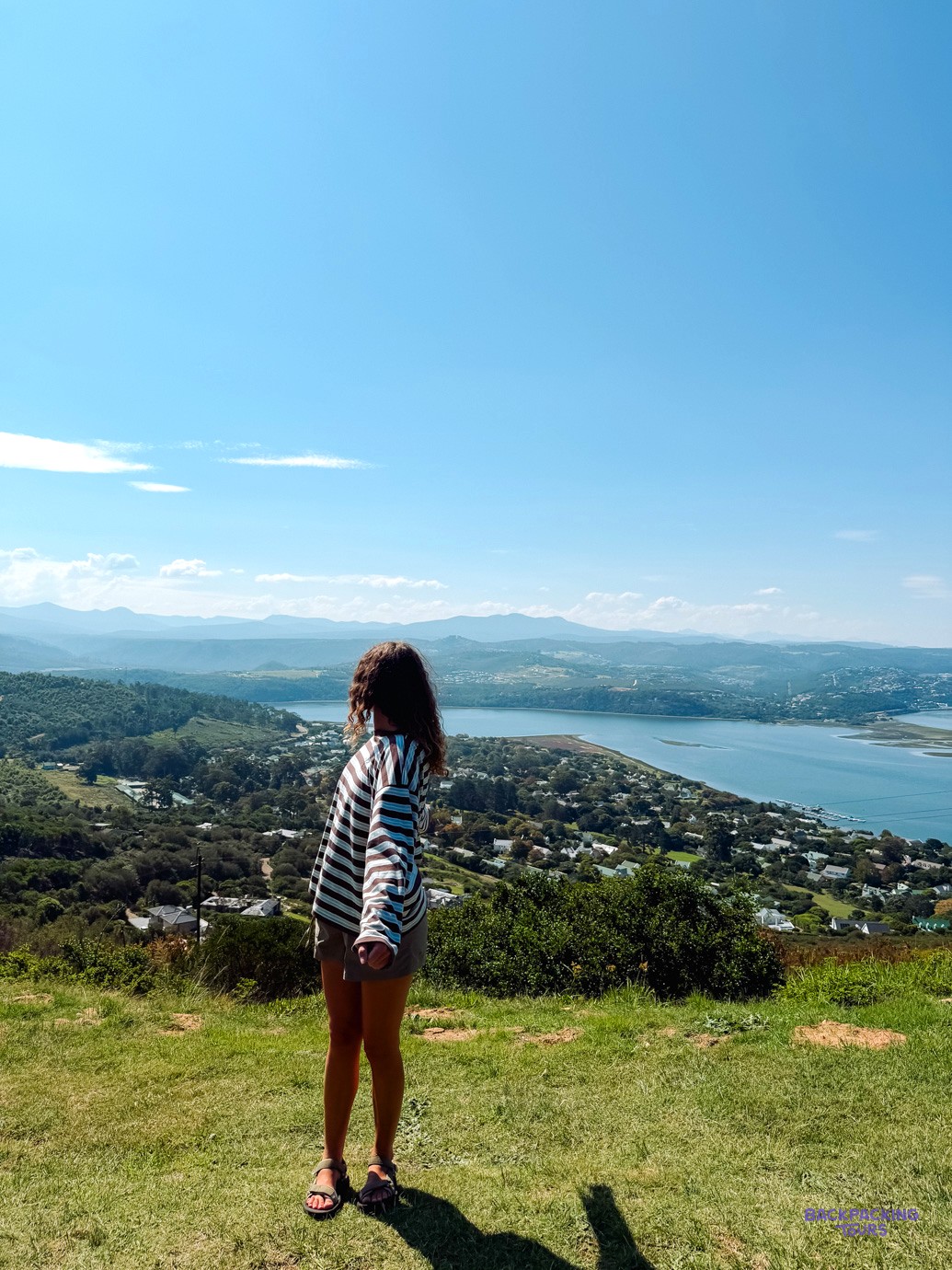 Traveller on a grassy hillside looking out over a wide lagoon and wooded town