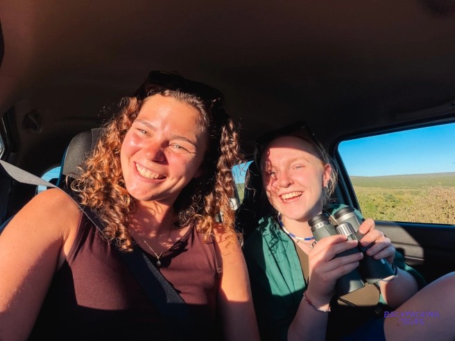 Two smiling travellers with binoculars riding in a car through open grassland