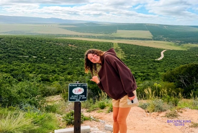 Traveller leaning by a beware of lions sign above a vast valley of green bush