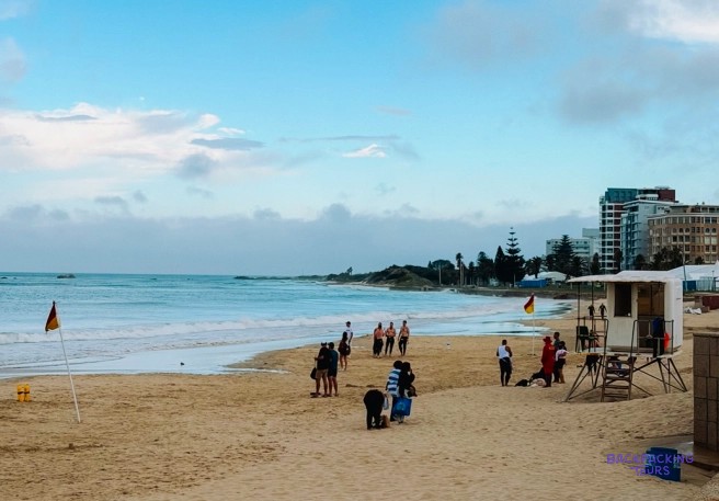 People strolling along a wide sandy beach past a lifeguard tower and safety flags