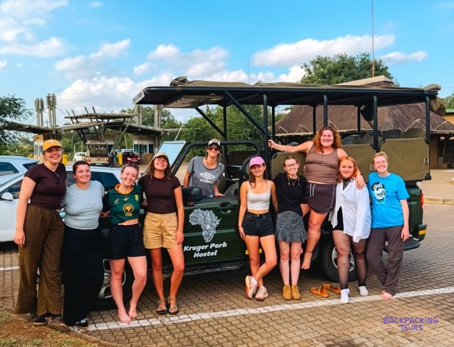 Group of travellers posing beside an open safari truck outside Kruger Park Hostel
