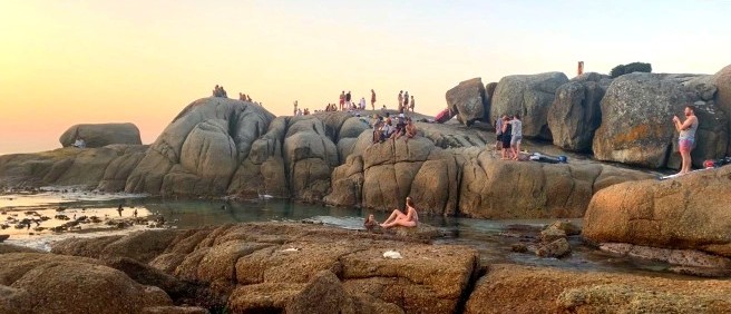 People sitting and climbing on huge granite boulders along the shore at sunset