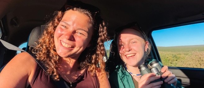 Two smiling travellers with binoculars riding in a car through open grassland
