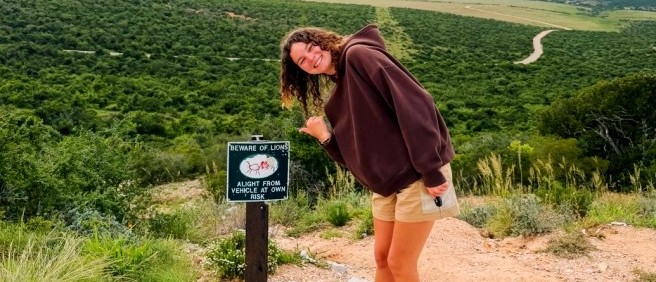 Traveller leaning by a beware of lions sign above a vast valley of green bush