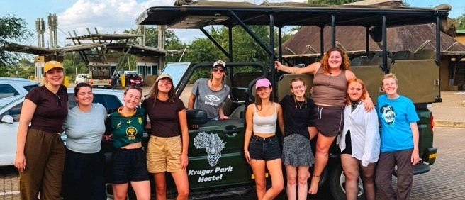 Group of travellers posing beside an open safari truck outside Kruger Park Hostel
