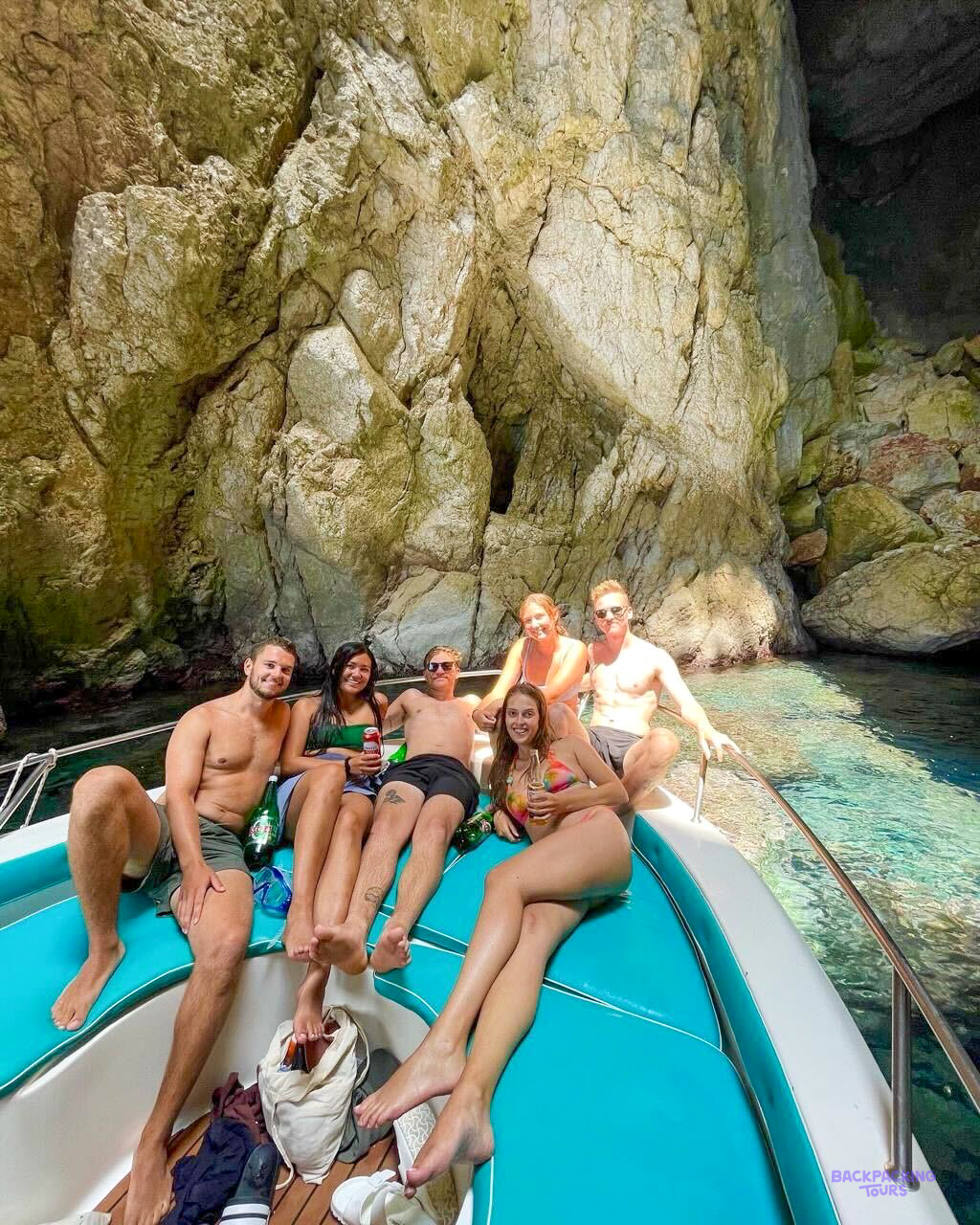 Six travellers relaxing on the bow of a boat in clear water beside the Himarë cliffs