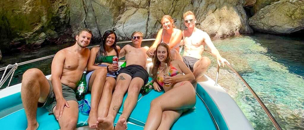 Six travellers relaxing on the bow of a boat in clear water beside the Himarë cliffs