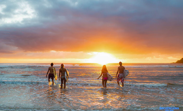 Backpackers carrying surfboards walk into the waves at a vibrant Byron Bay sunset.