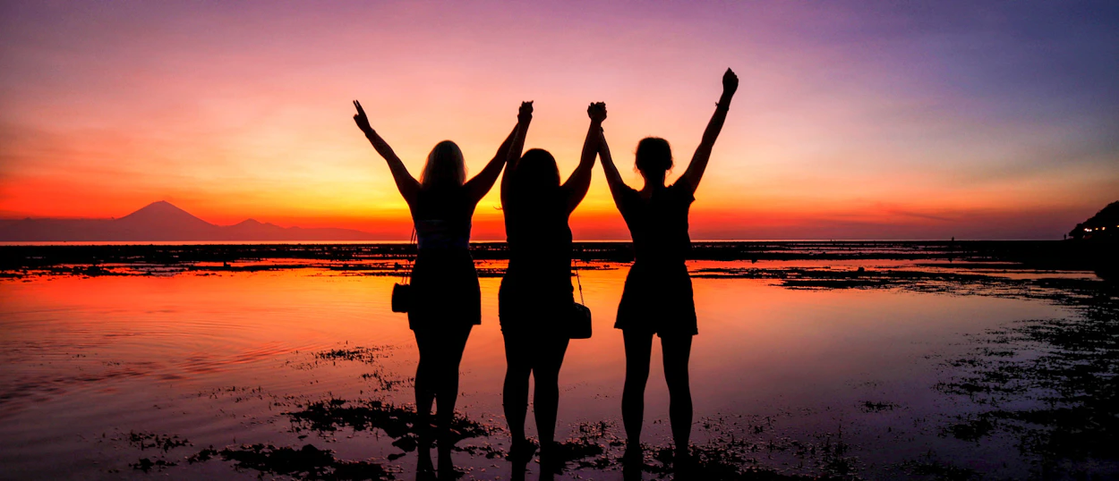 Three friends celebrate on the beach at sunset, arms raised in joy on Gili Trawangan.