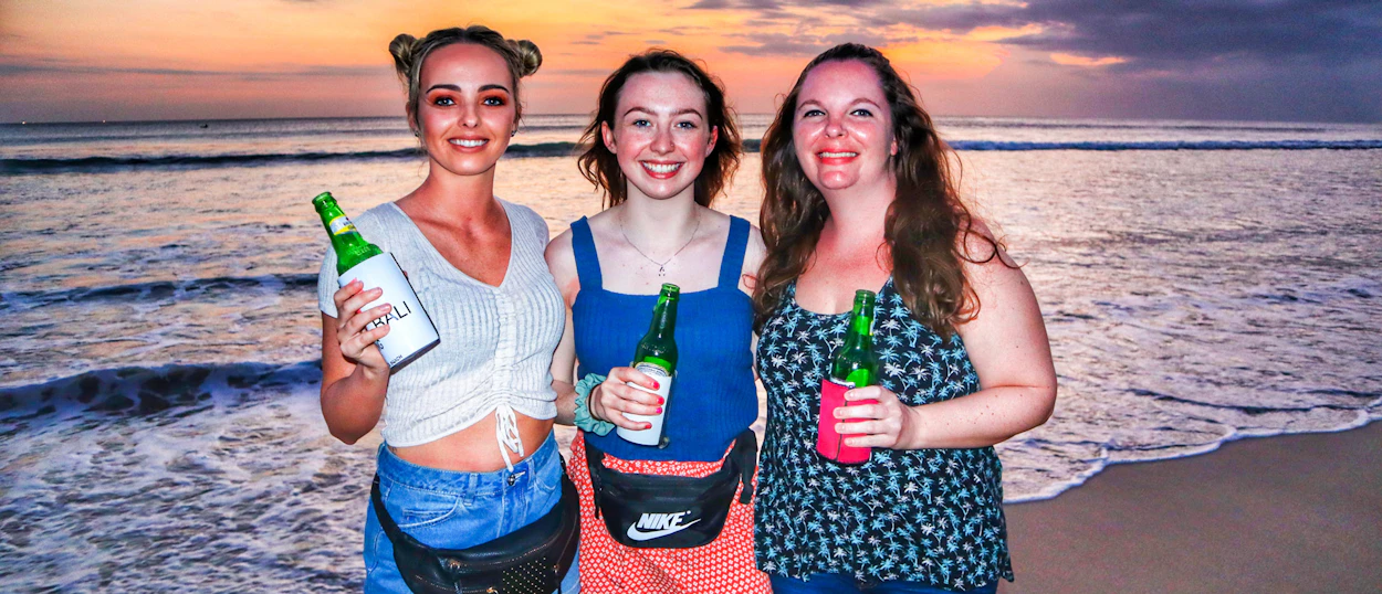 Three happy women holding drinks on a beach at sunset in Canggu, Bali.