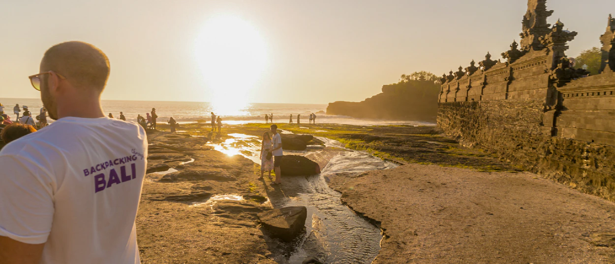 A backpacker watches the golden sunset over a rocky Bali beach with an ornate temple wall.