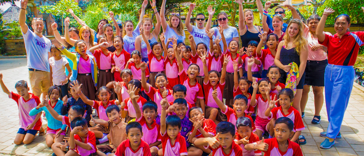 A large group of smiling children and adults enthusiastically wave their hands at a school in Ubud, Bali.