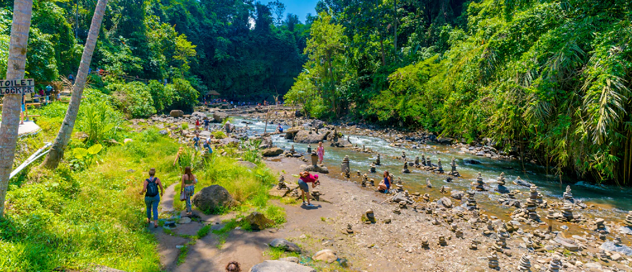 Travelers explore a rocky riverbed with stacked stones amidst lush green jungle in Ubud, Bali.