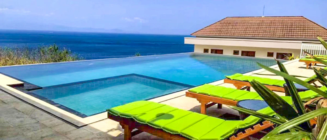 An infinity pool with vibrant green lounge chairs overlooks the blue ocean on Nusa Penida.