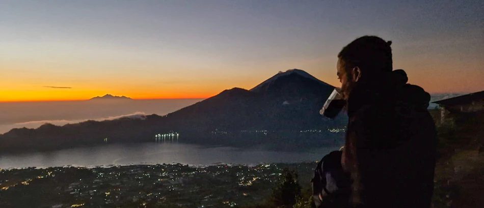 A person sips coffee at sunrise from Mount Batur summit, overlooking Lake Batur and volcanic landscape.