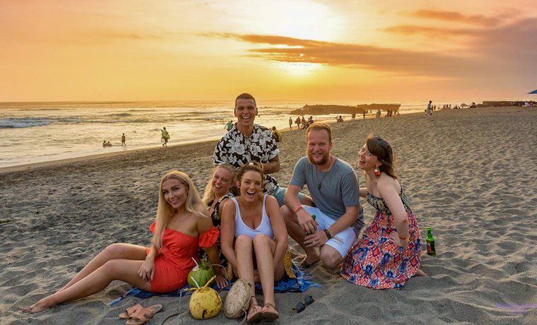Six friends smiling and relaxing on Canggu beach at sunset, with coconuts on the sand.
