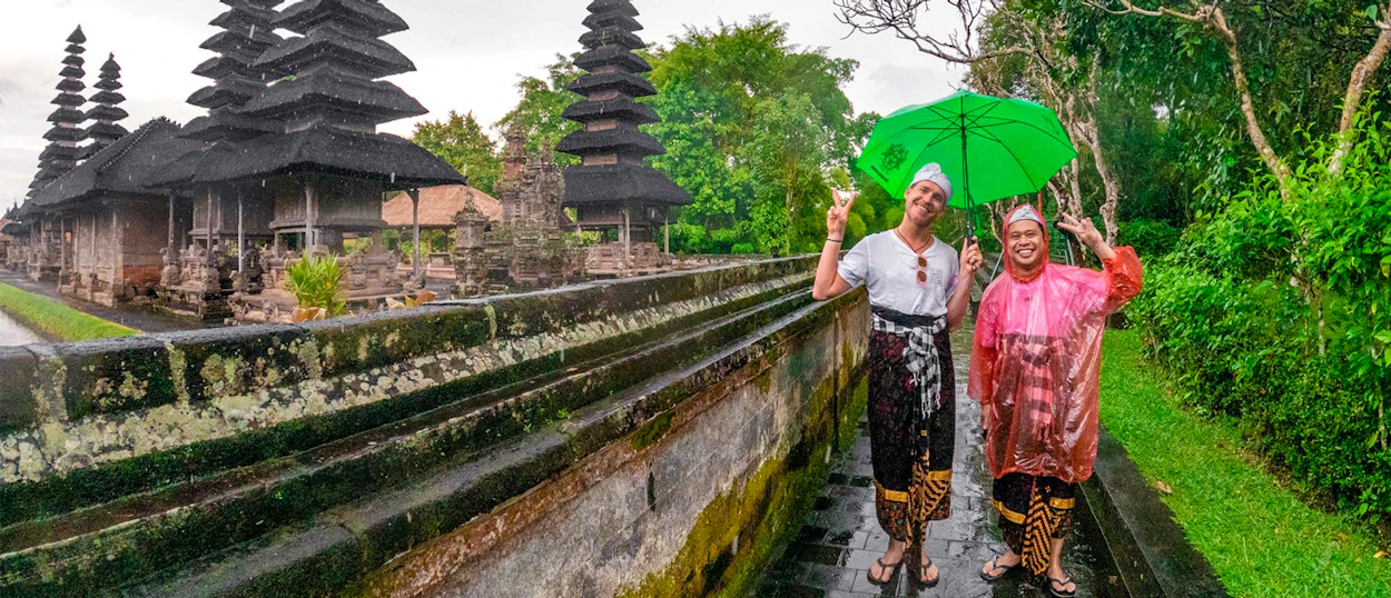 Two smiling people in traditional Balinese sarongs visit an ancient temple in Canggu during rain.