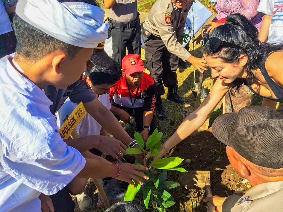 A group of people, including locals and a tourist, collaboratively plant a small tree in Bali.