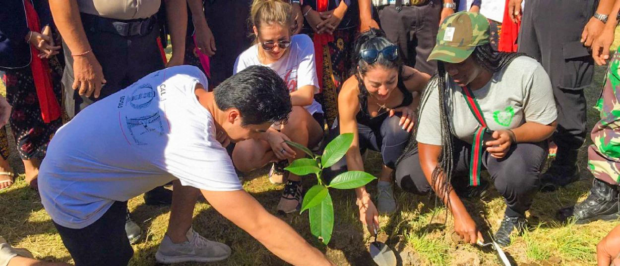 Young adults collaboratively planting a small tree in a grassy area, likely during an environmental program in Bali.