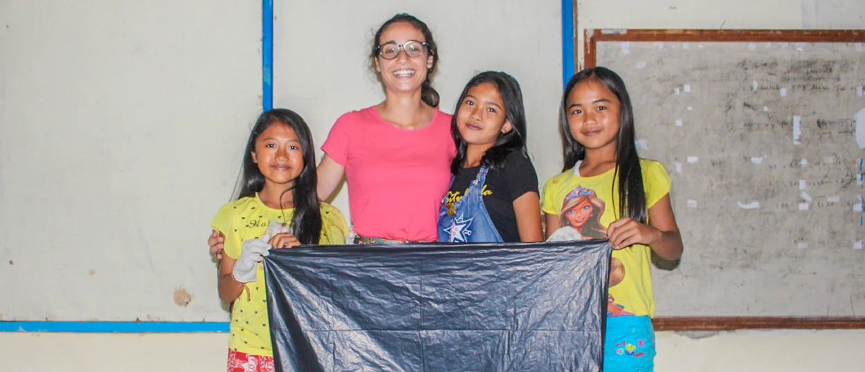 A smiling woman and three Balinese primary school girls holding a black bag in a classroom.