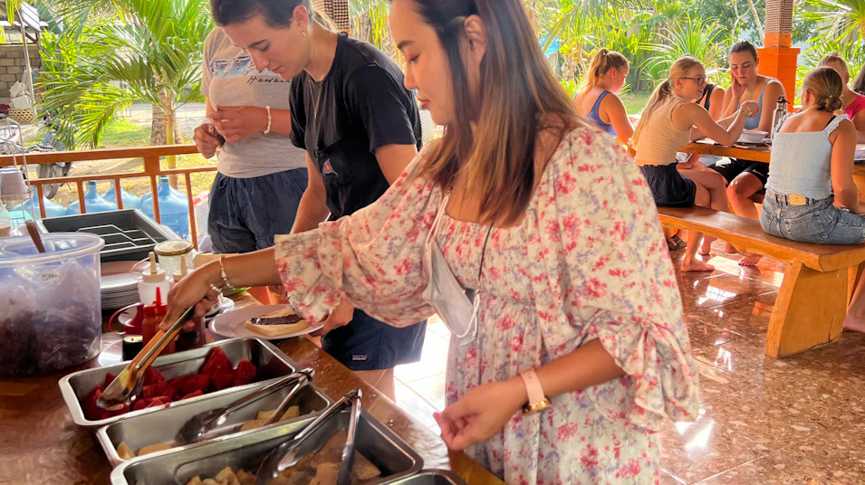 Young travelers serving themselves a casual buffet meal in a bright, tropical outdoor setting.