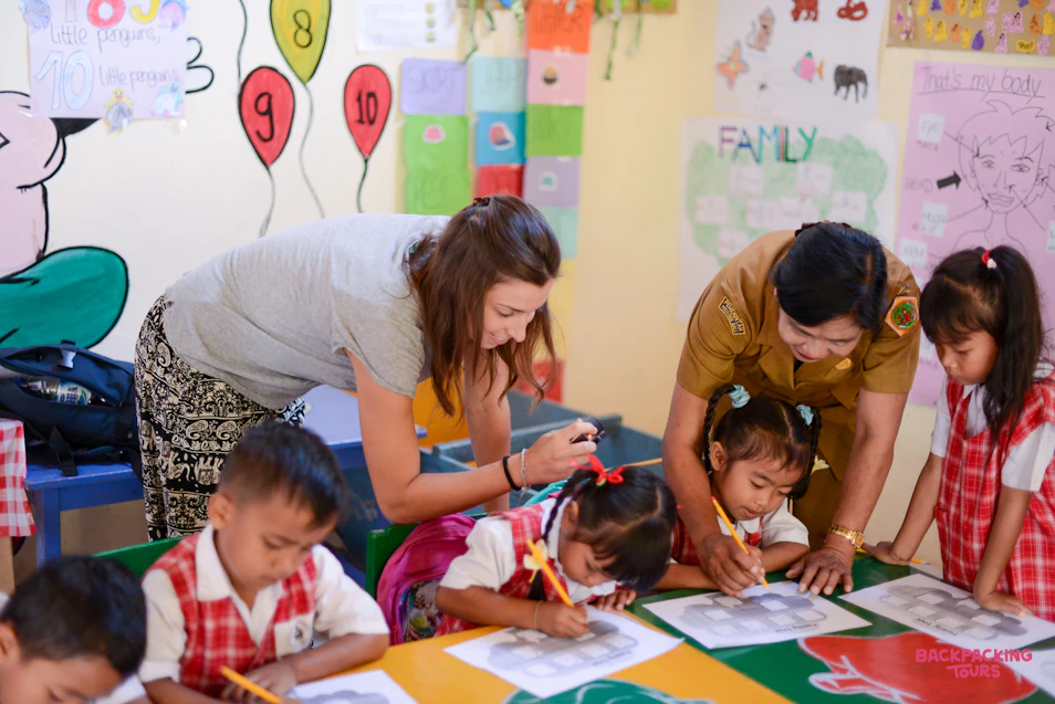 A volunteer and teacher help young children draw and learn in a colorful Bali kindergarten classroom.