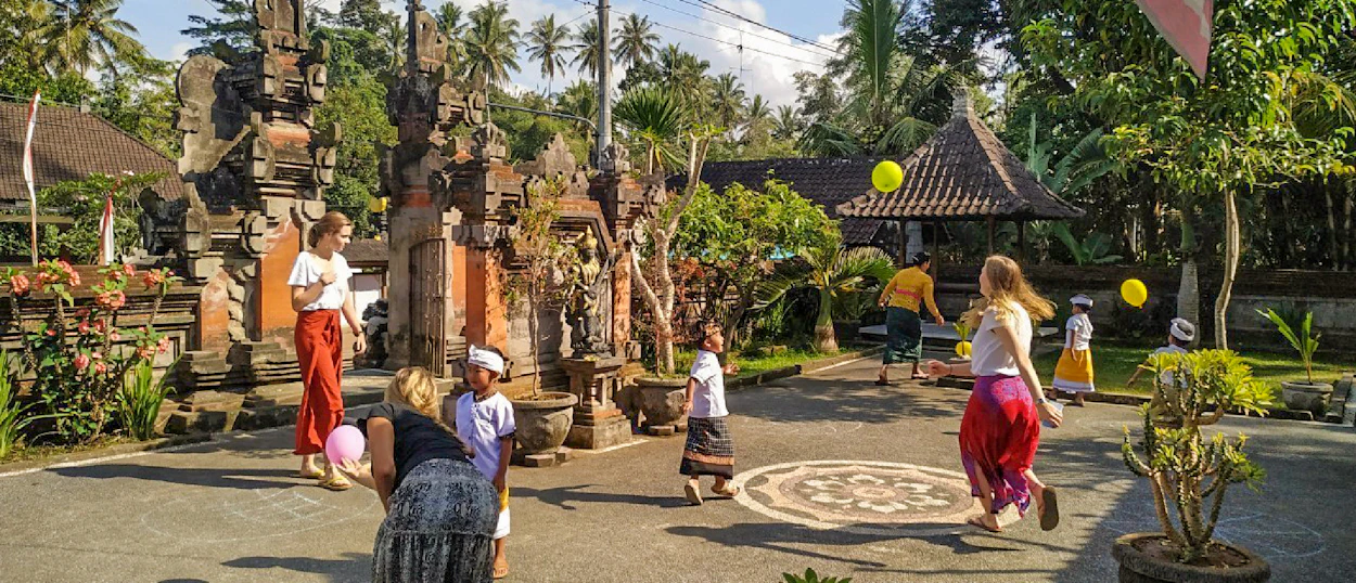 Adults and Balinese children joyfully play with balloons in a sunny temple courtyard in Bali.