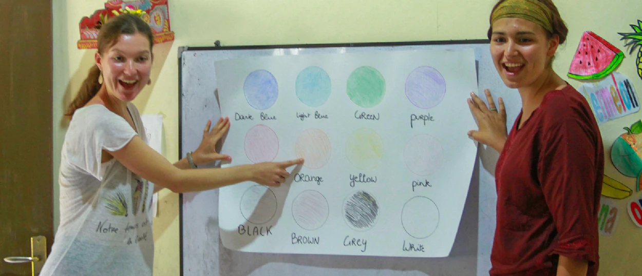 Two enthusiastic women teaching colors from a poster in a bright kindergarten classroom in Bali.