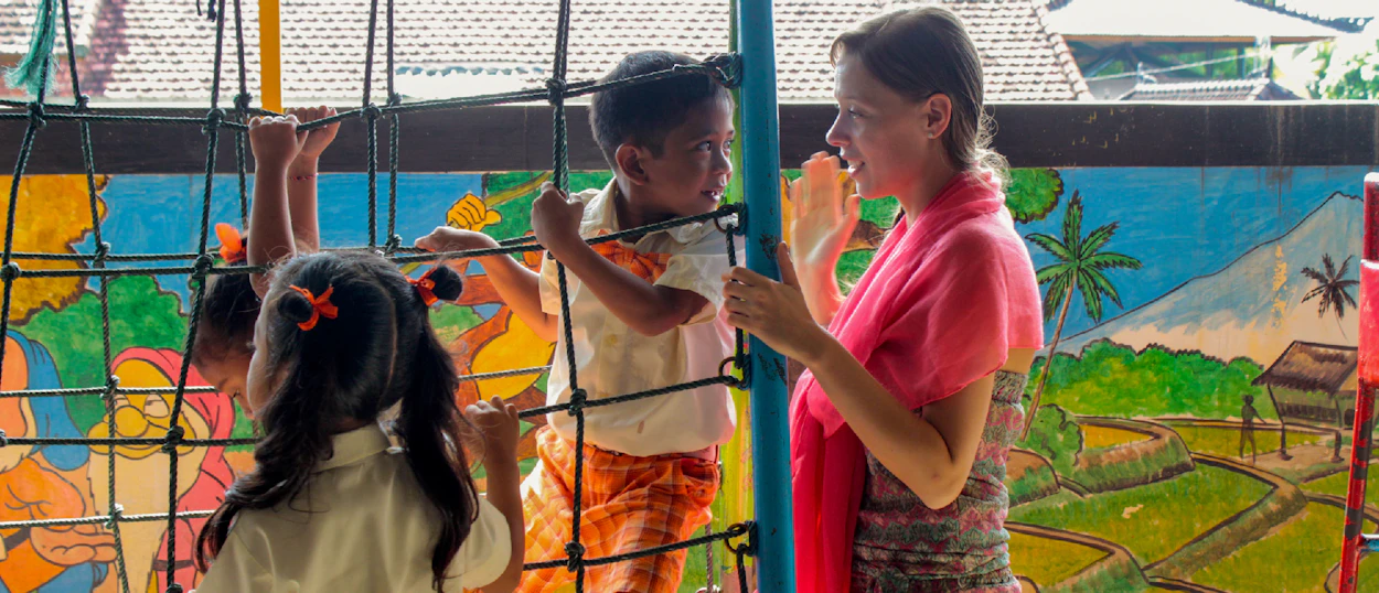 A volunteer teacher engages with children playing on a climbing net at a kindergarten in Bali.