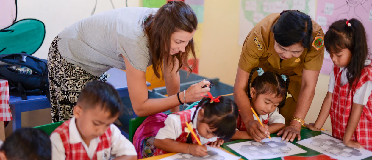 Volunteer and local teacher helping young children draw and learn in a bright classroom in Bali.