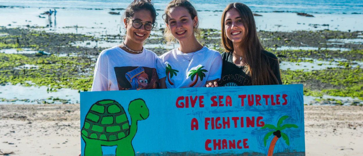 Smiling women hold a turtle conservation sign on a sandy beach with shallow water in Nusa Penida.