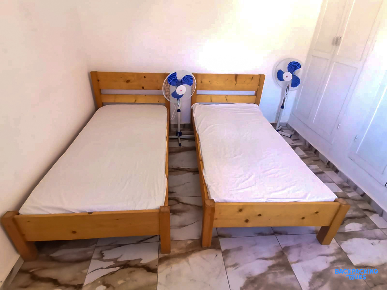 A simple room featuring two single wooden beds with white sheets and two electric fans.
