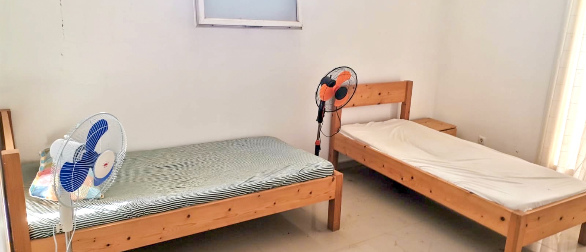 A basic shared room featuring two single wooden beds and standing fans.