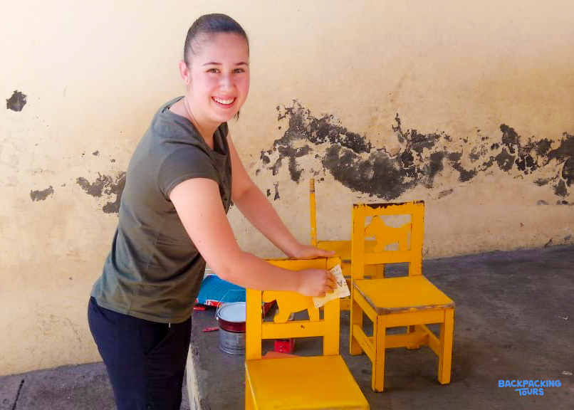 A smiling volunteer paints small yellow chairs, contributing to a renovation project.