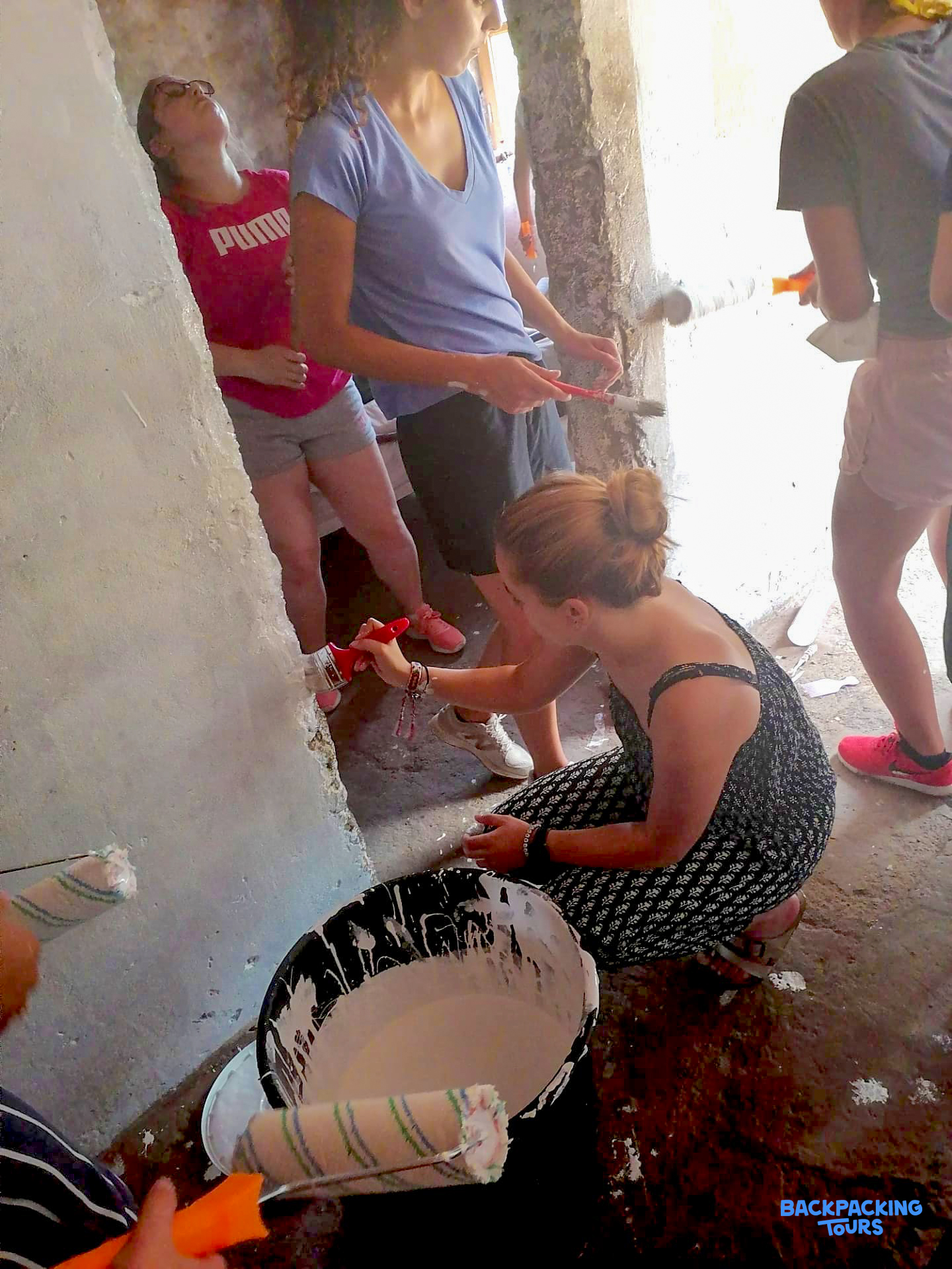 A group of young volunteers actively paint a rough interior wall white with rollers and brushes.