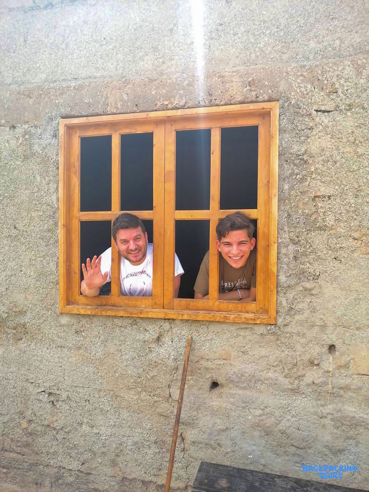 Two smiling men wave from a wooden window in a rustic wall, looking friendly and welcoming.
