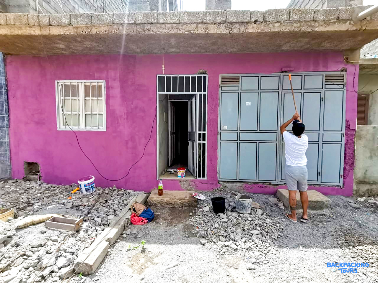 A person paints the bright pink exterior of a building with a roller, surrounded by renovation materials.
