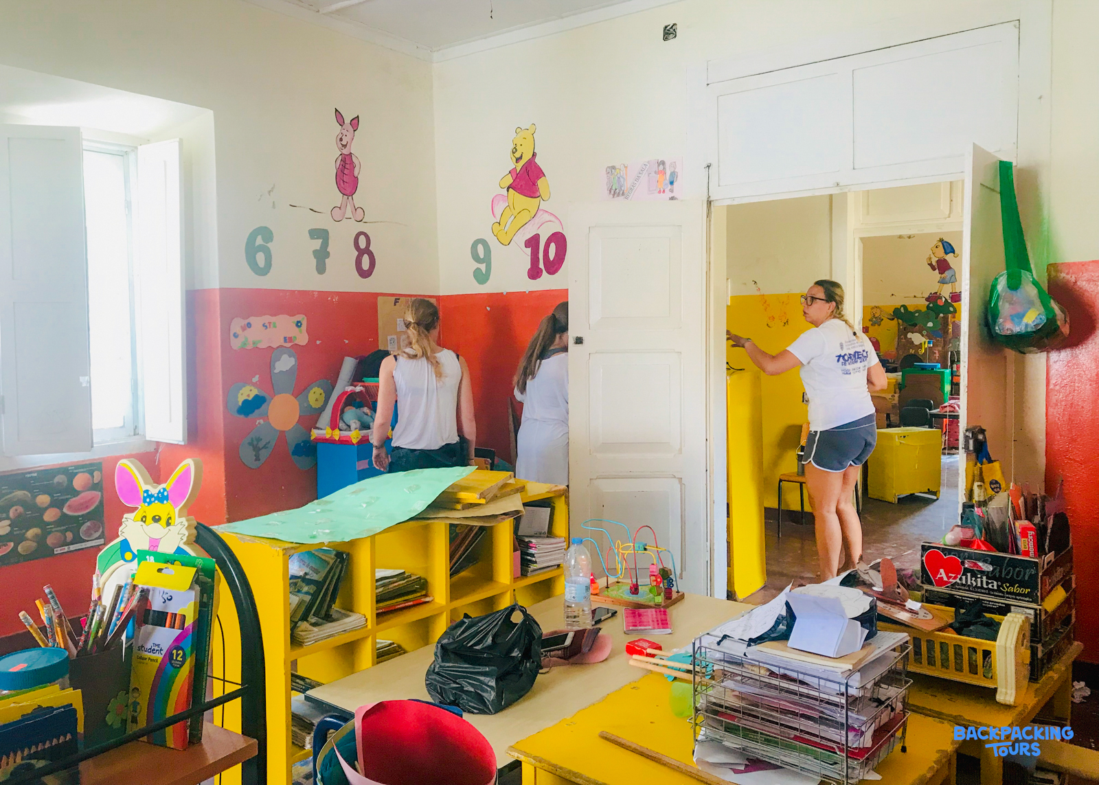 Volunteers brighten a classroom, painting a door frame and organizing colorful desks with children's art supplies.