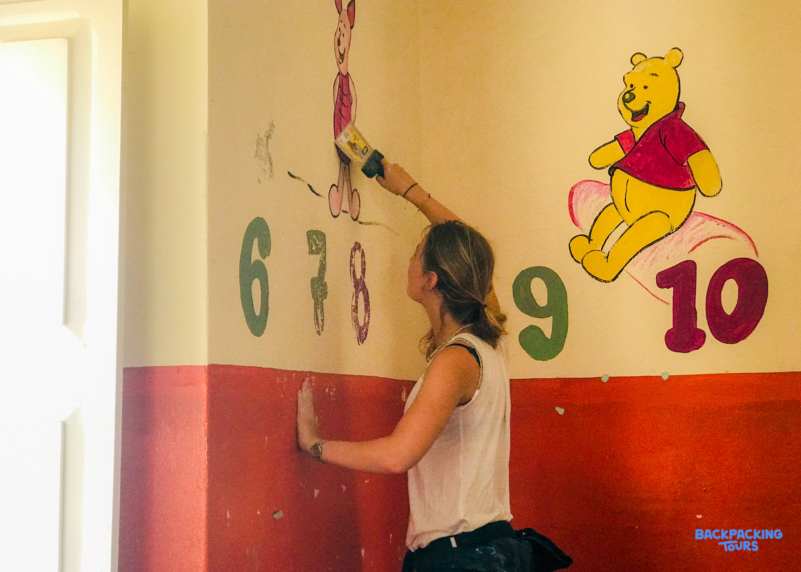 A woman paints a colorful wall mural featuring cartoon characters and numbers in a bright room.