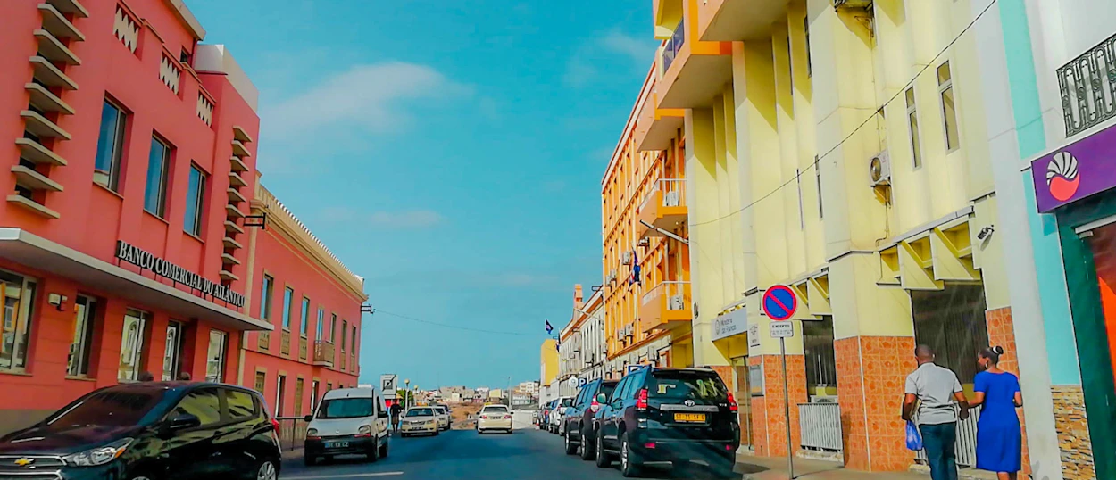Two people walk past colorful buildings and parked cars on a sunny street.
