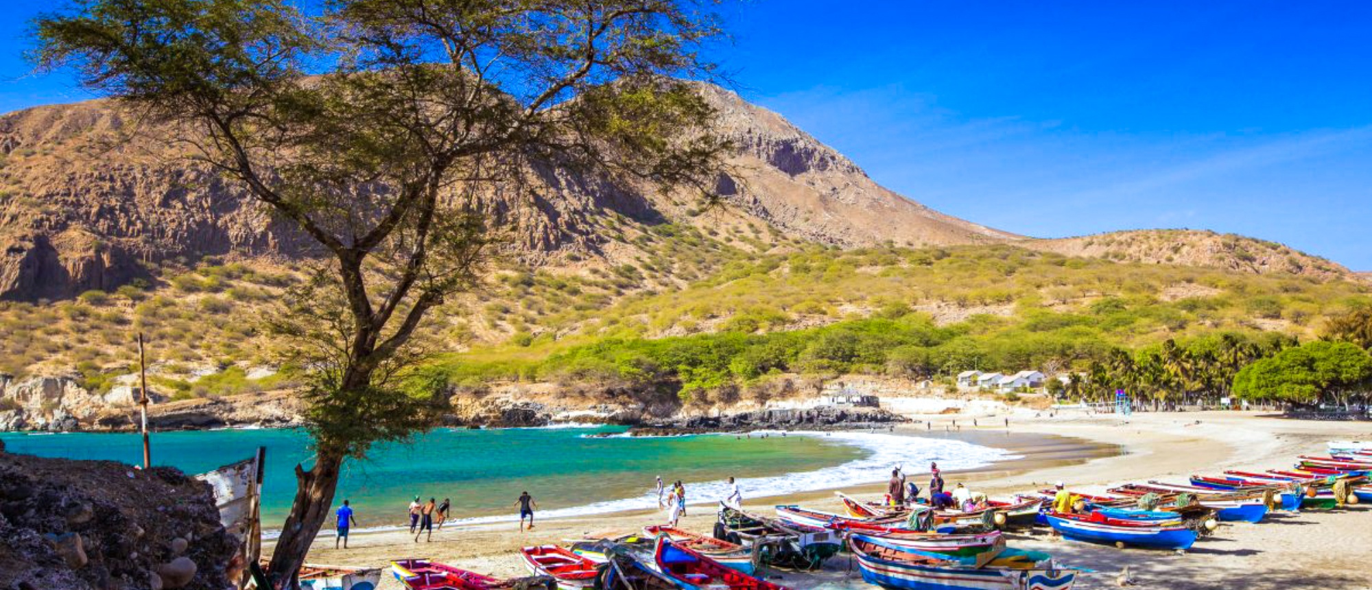 Colorful fishing boats line a sunny beach with clear turquoise water and arid mountains in Cape Verde.