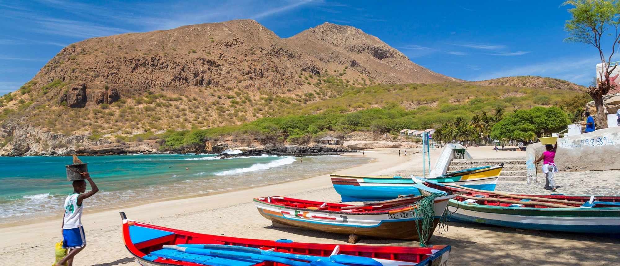 Colorful fishing boats on a sunny beach with people carrying baskets, backed by arid mountains.