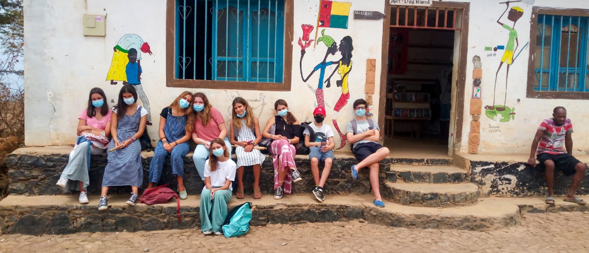 A group of masked volunteers sit on steps in front of a building with colorful murals.