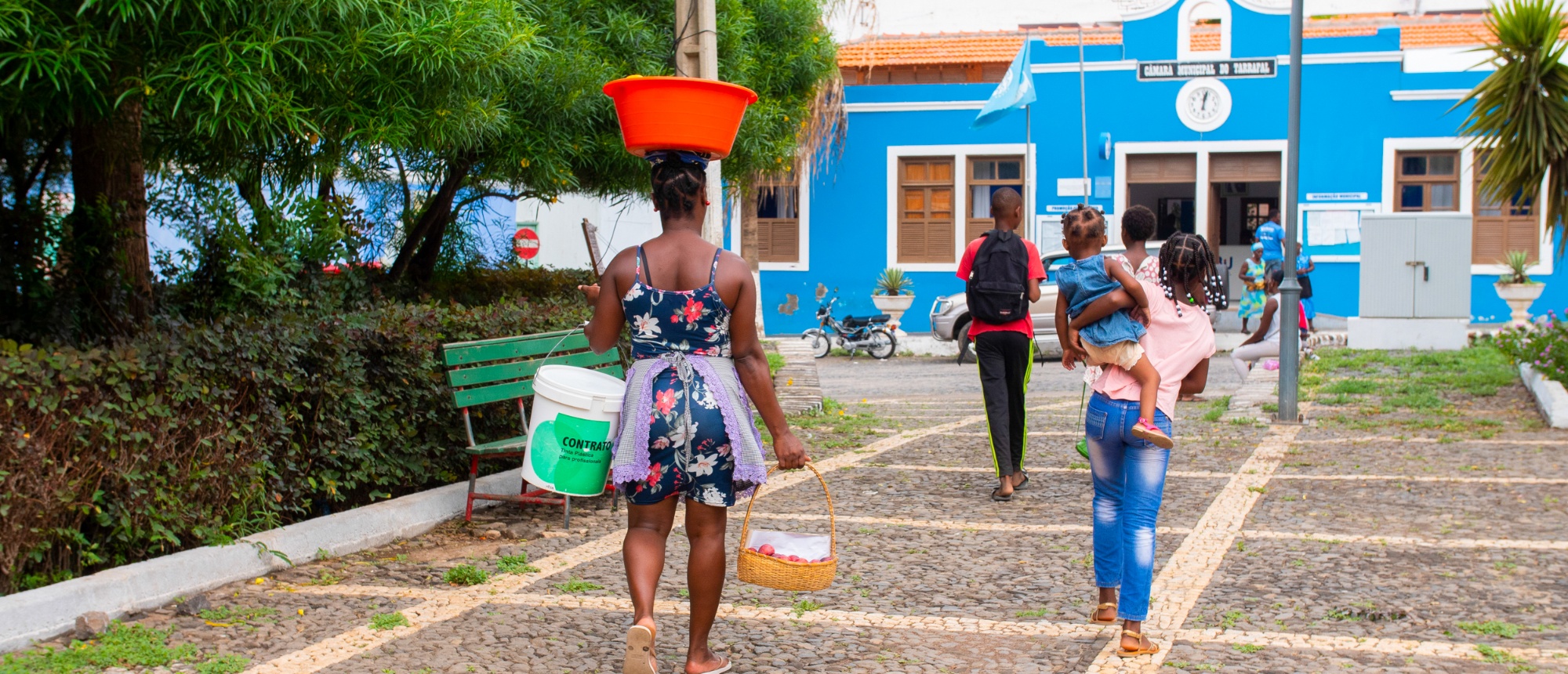 People walk on a sunny paved path, passing a vibrant blue building in Cape Verde.