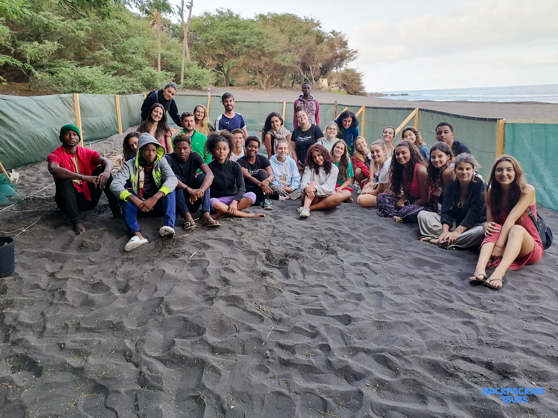A large group of diverse young travelers sitting together on a dark sand beach.